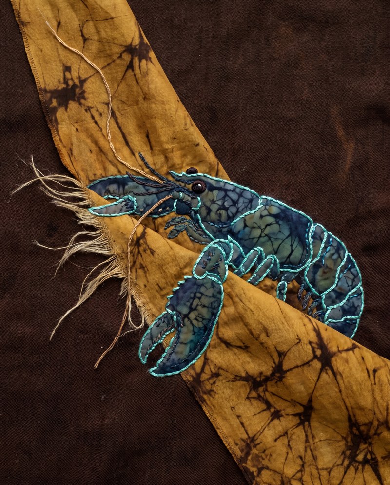 The Useful Roughness — tactile batik and raised silk stitching in ochre-gold and espresso-brown: a small blue-green creature reclining along the steep diagonal fold of a suspended silk ribbon whose raw edge is still visibly separated into pale flax fibers.